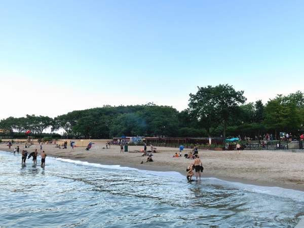 Small groups of people on a quiet lakeside beach at dusk, with low green trees behind them