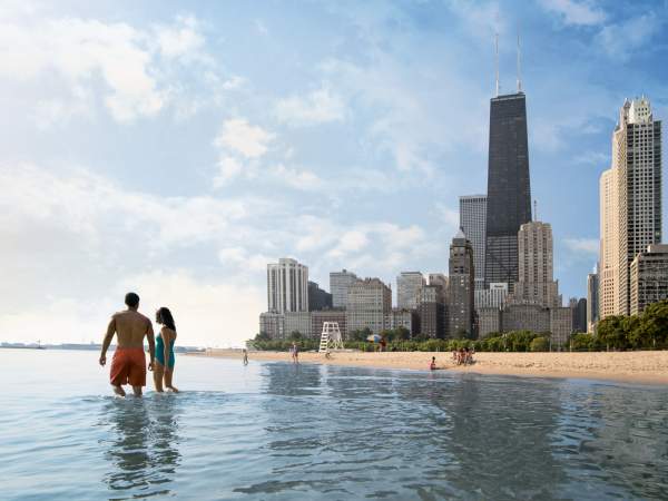 A couple walking through the shallows of Lake Michigan next to a sandy shore, with skyscrapers in the background